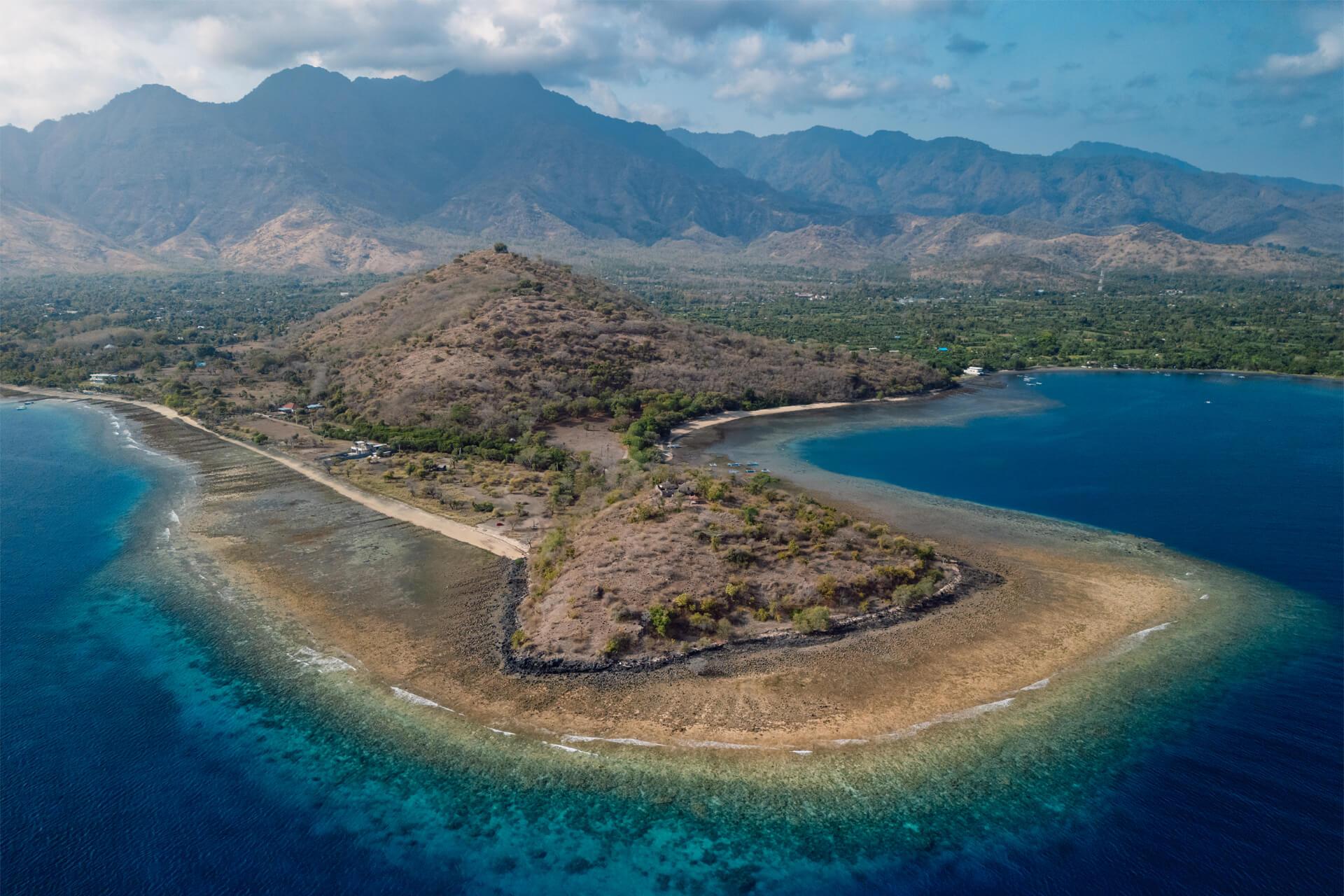 Plage de Pemuteran entourée d’eau turquoise et de récifs coralliens, avec collines arides et montagnes en arrière-plan.