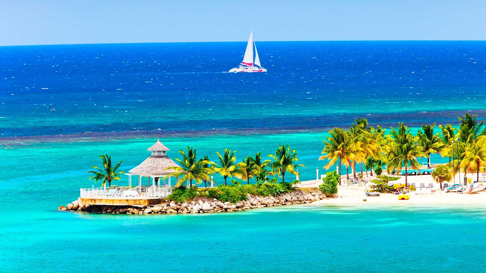 Plage d’Ocho Rios en Jamaïque, avec sable blanc, palmiers, eau turquoise de la mer des Caraïbes et un voilier blanc au loin.