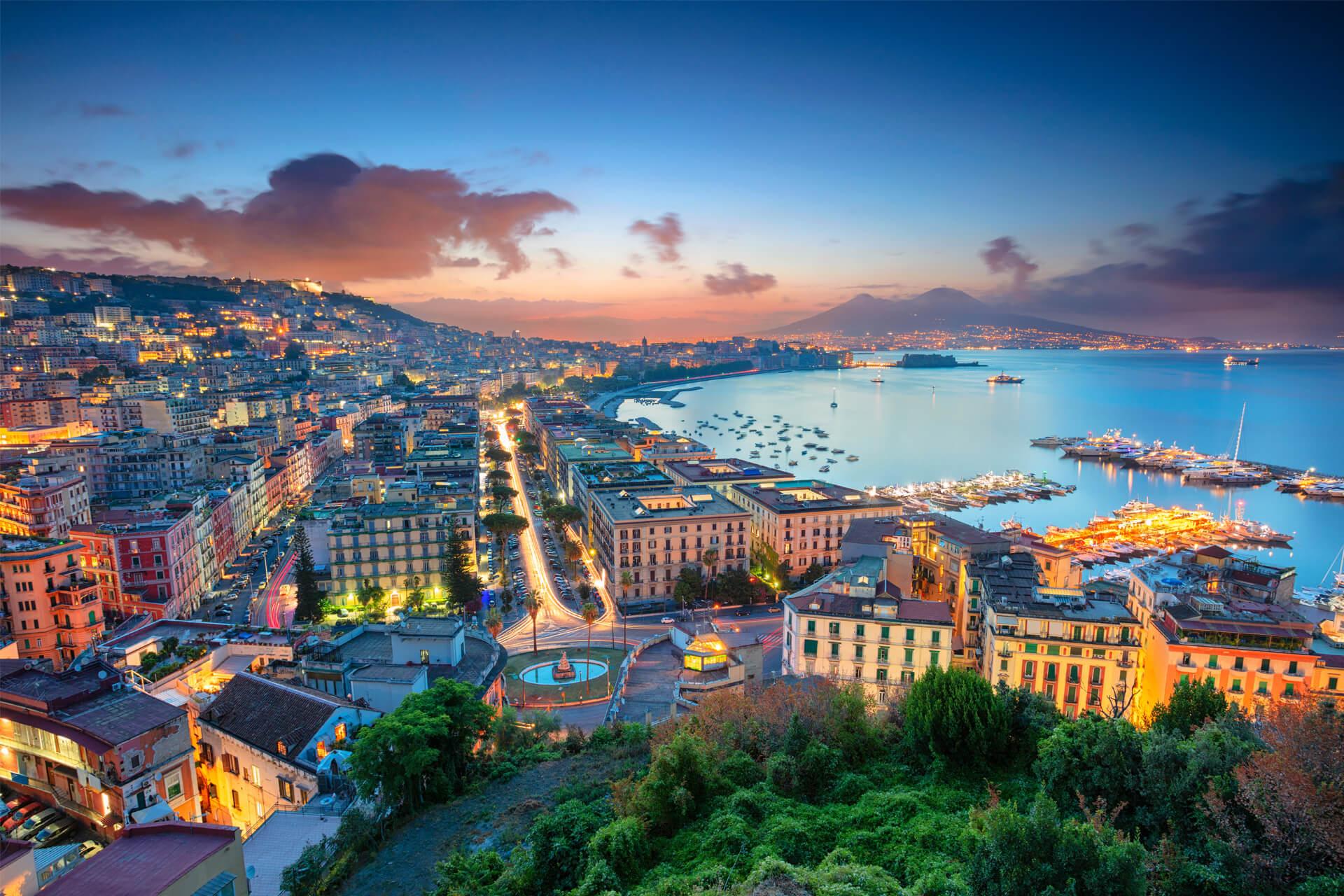 Panoramic view of Naples at sunset, featuring its sparkling lights, bustling harbor, and Mount Vesuvius in the background.