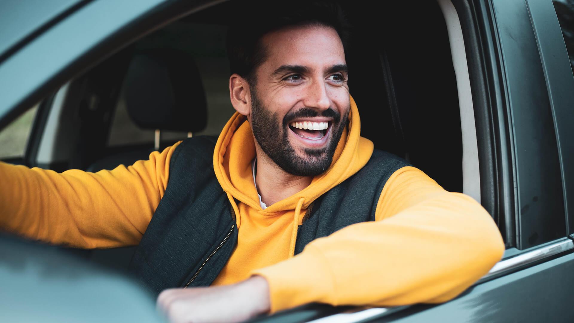 Gros plan sur une femme qui, assise dans sa voiture, regarde par la fenêtre vers l’arrière, une main sur le volant et l’autre appuyée sur le rebord de la fenêtre.