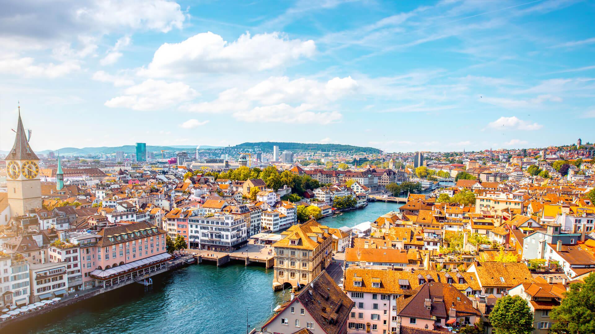Vue panoramique de Zurich en Suisse, avec la rivière Limmat, toits en tuiles rouges, clochers historiques et collines verdoyantes.