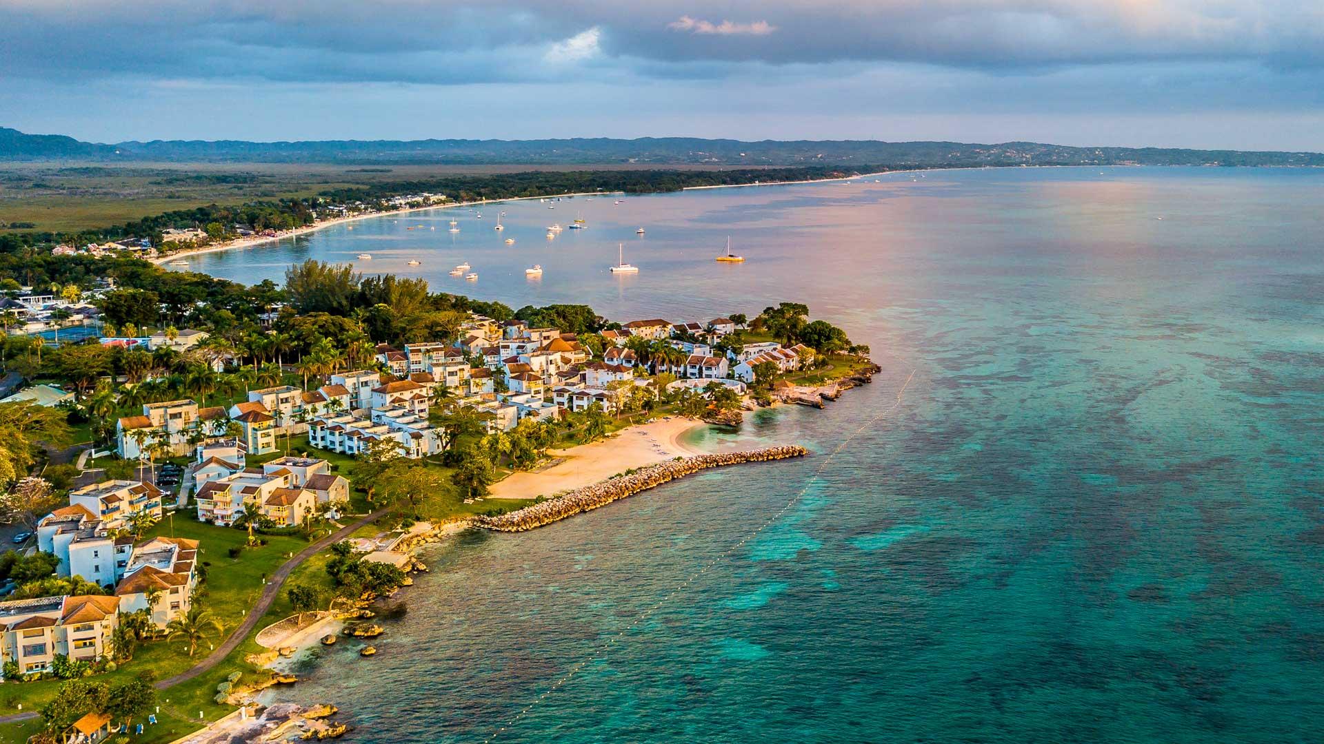 Aerial view of Negril, Jamaica, showcasing turquoise waters, lush beachfront vegetation, and white seaside buildings.
