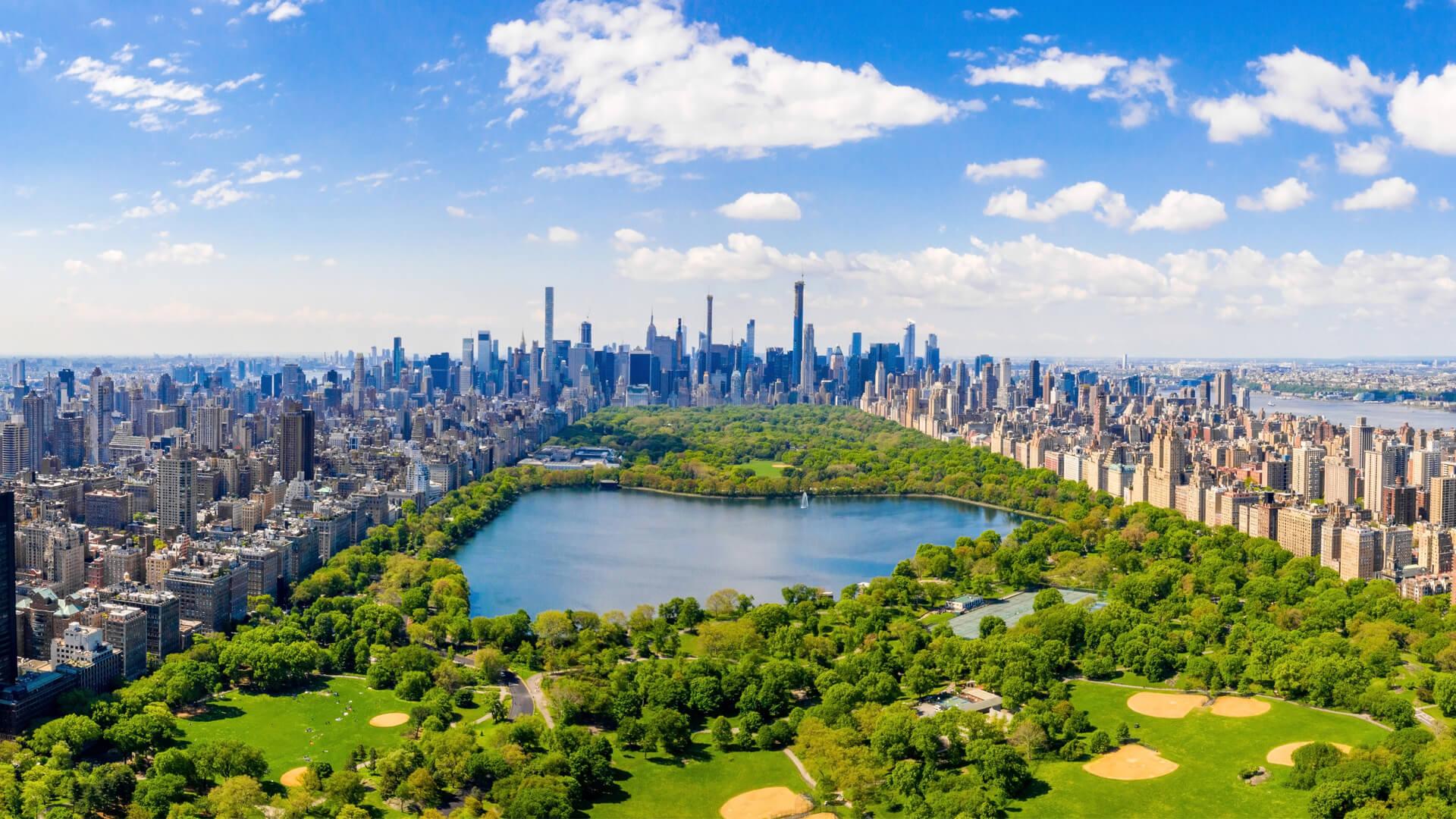 Aerial view of Central Park in New York, featuring the lake, lush greenery, Manhattan skyscrapers, and a clear blue sky.