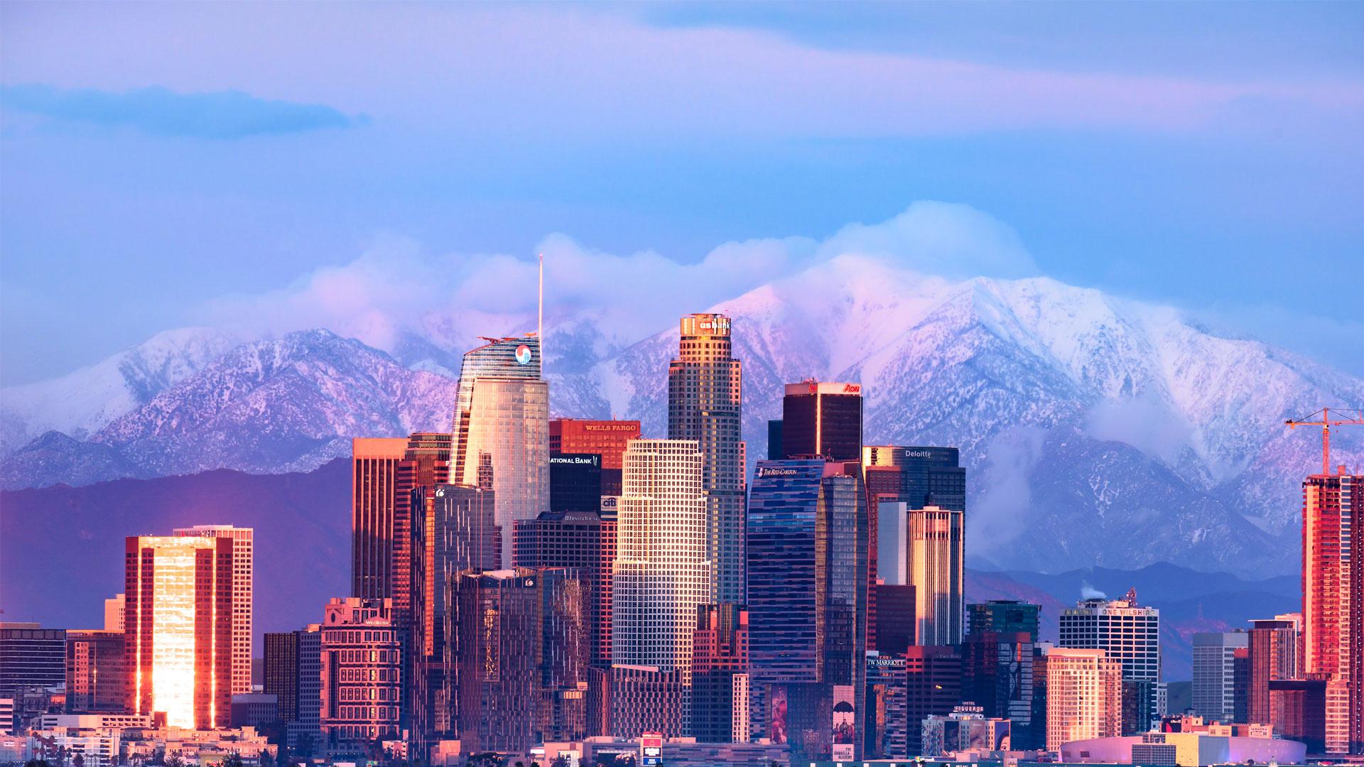 Gratte-ciel de Los Angeles baignés de teintes roses et dorées au coucher du soleil, avec montagnes enneigées en arrière-plan.