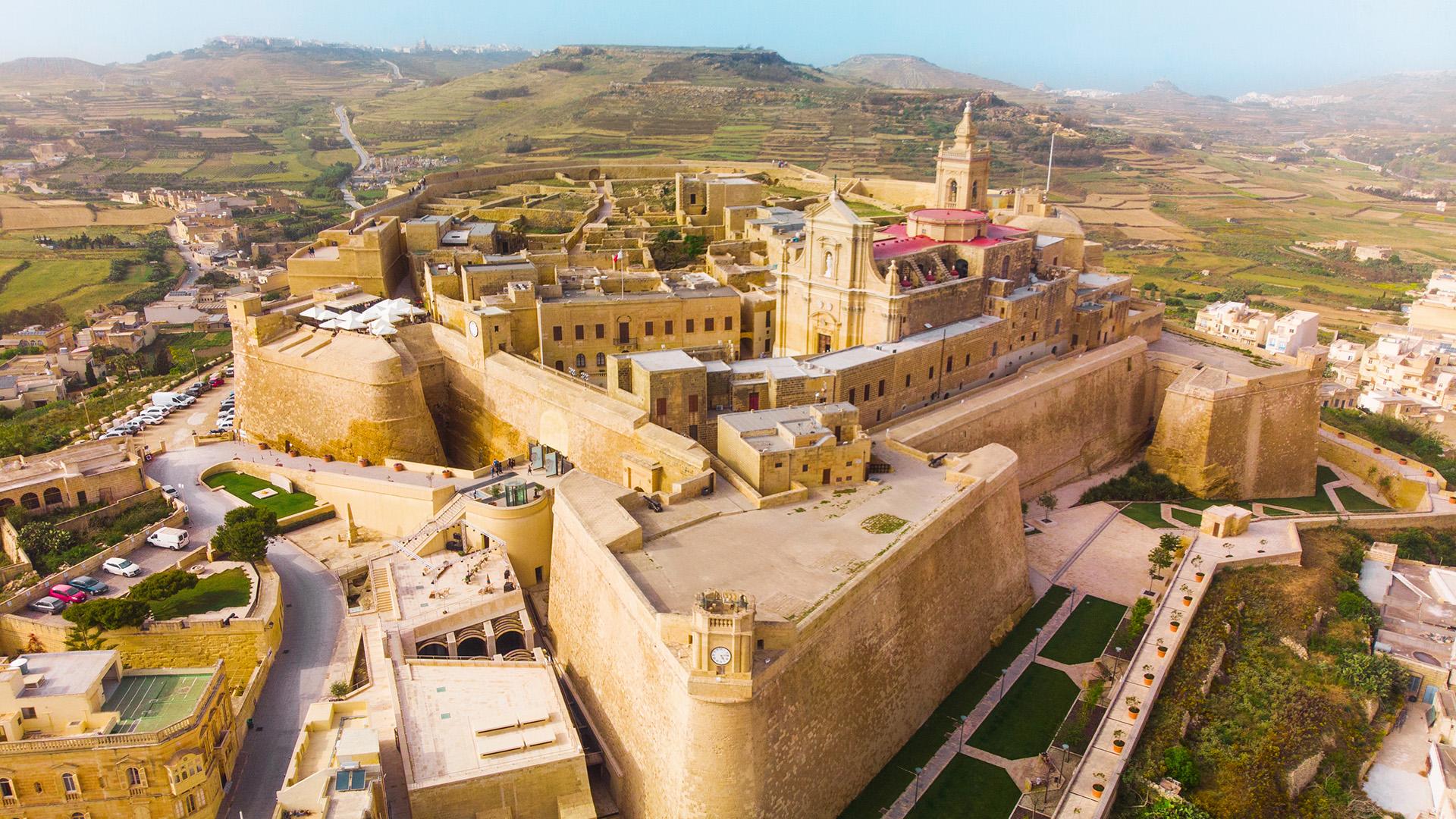Vue aérienne de la ville de Rabat à Gozo, avec citadelle fortifiée, église baroque et campagne maltaise en arrière-plan.