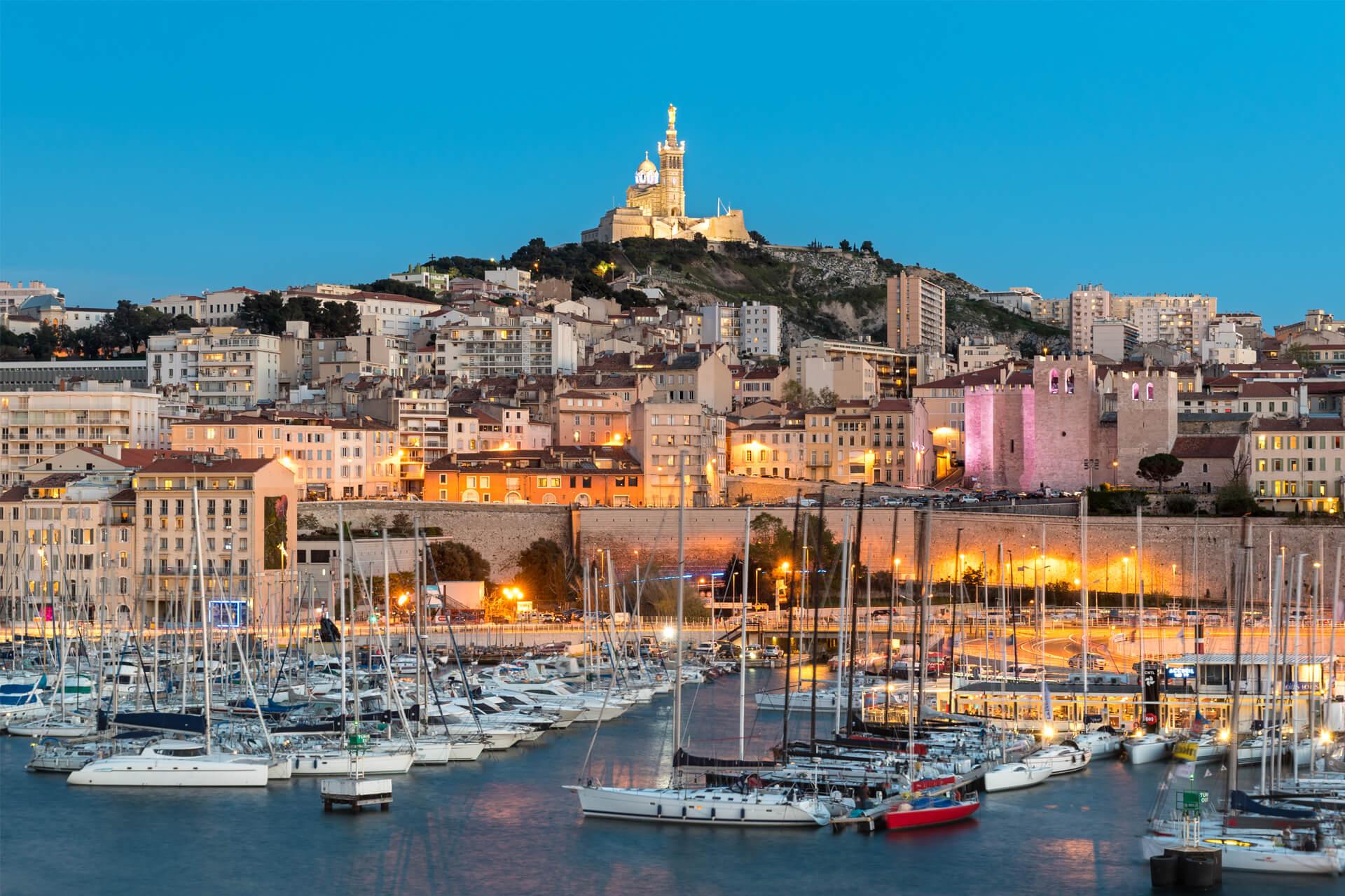Marseille’s Old Port at twilight with docked sailboats, glowing ramparts, and the basilica overlooking the city.