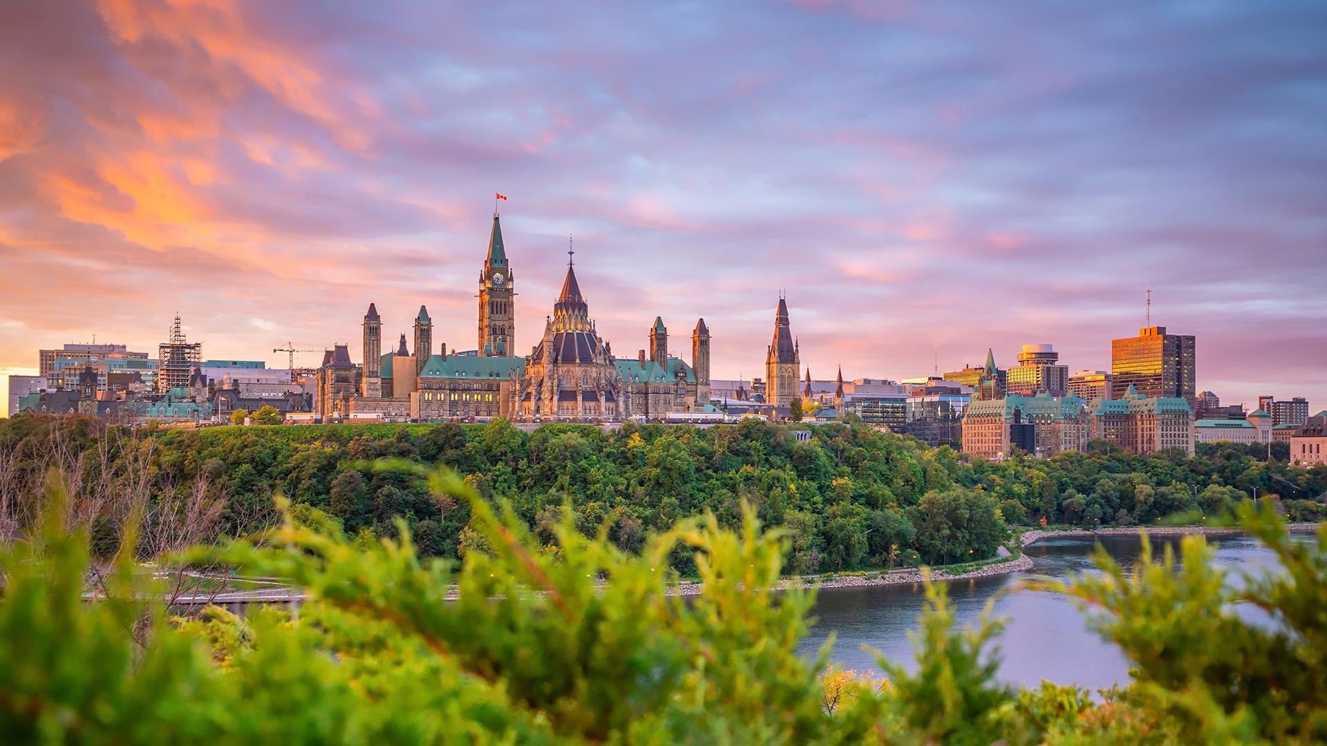 Vue panoramique d’Ottawa et de la rivière des Outaouais au coucher du soleil, avec le Parlement du Canada dominant la ville.