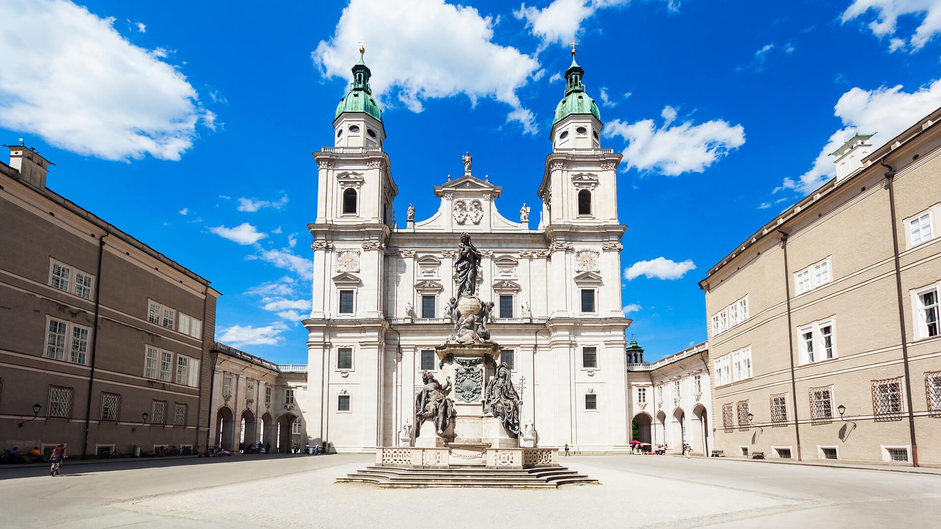 Facade of Salzburg Cathedral in Austria under a blue sky, with a statue in the foreground.