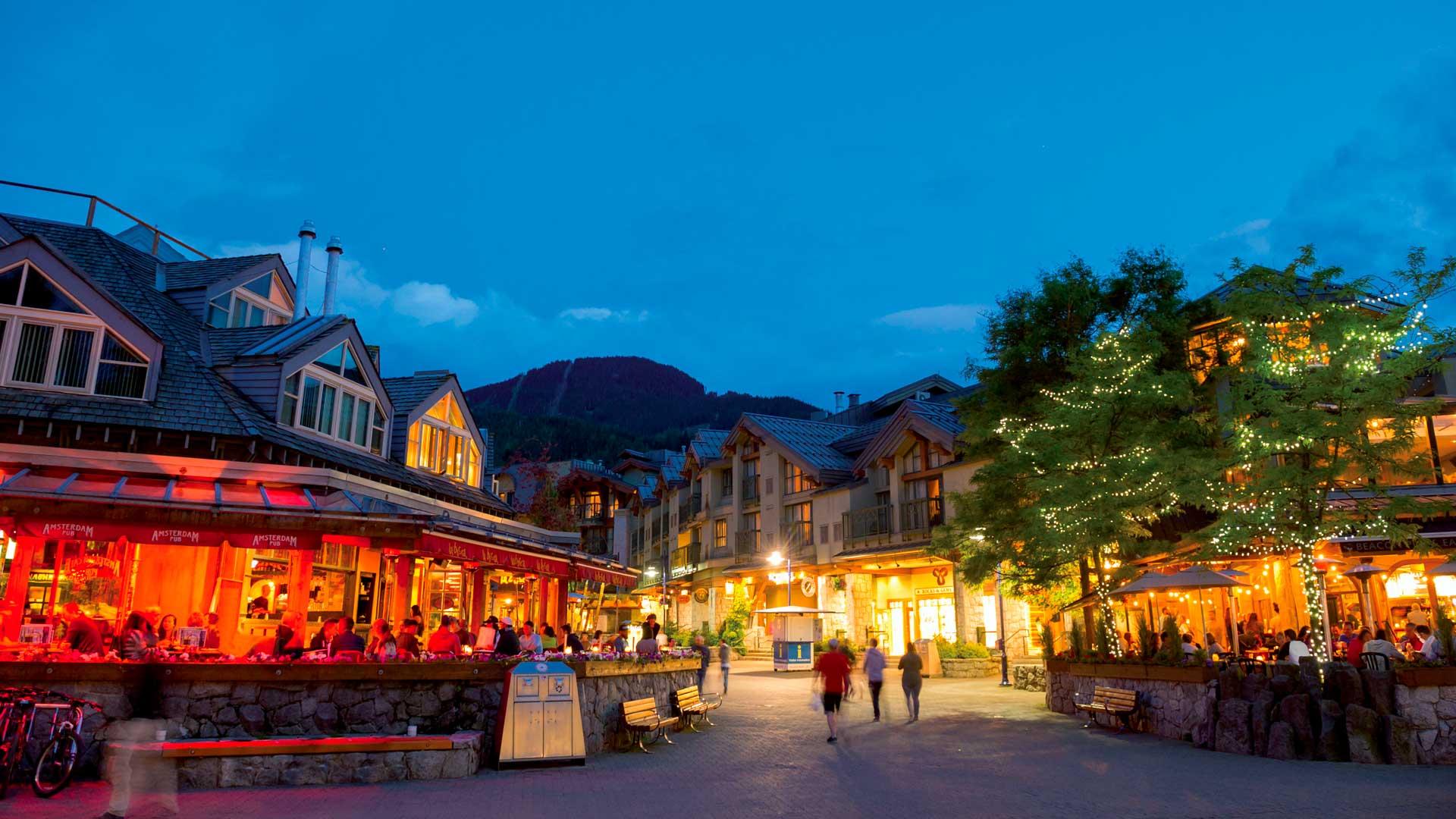 Village piétonnier de Whistler au crépuscule, avec chalets alpins éclairés, restaurants animés et montagnes en arrière-plan.