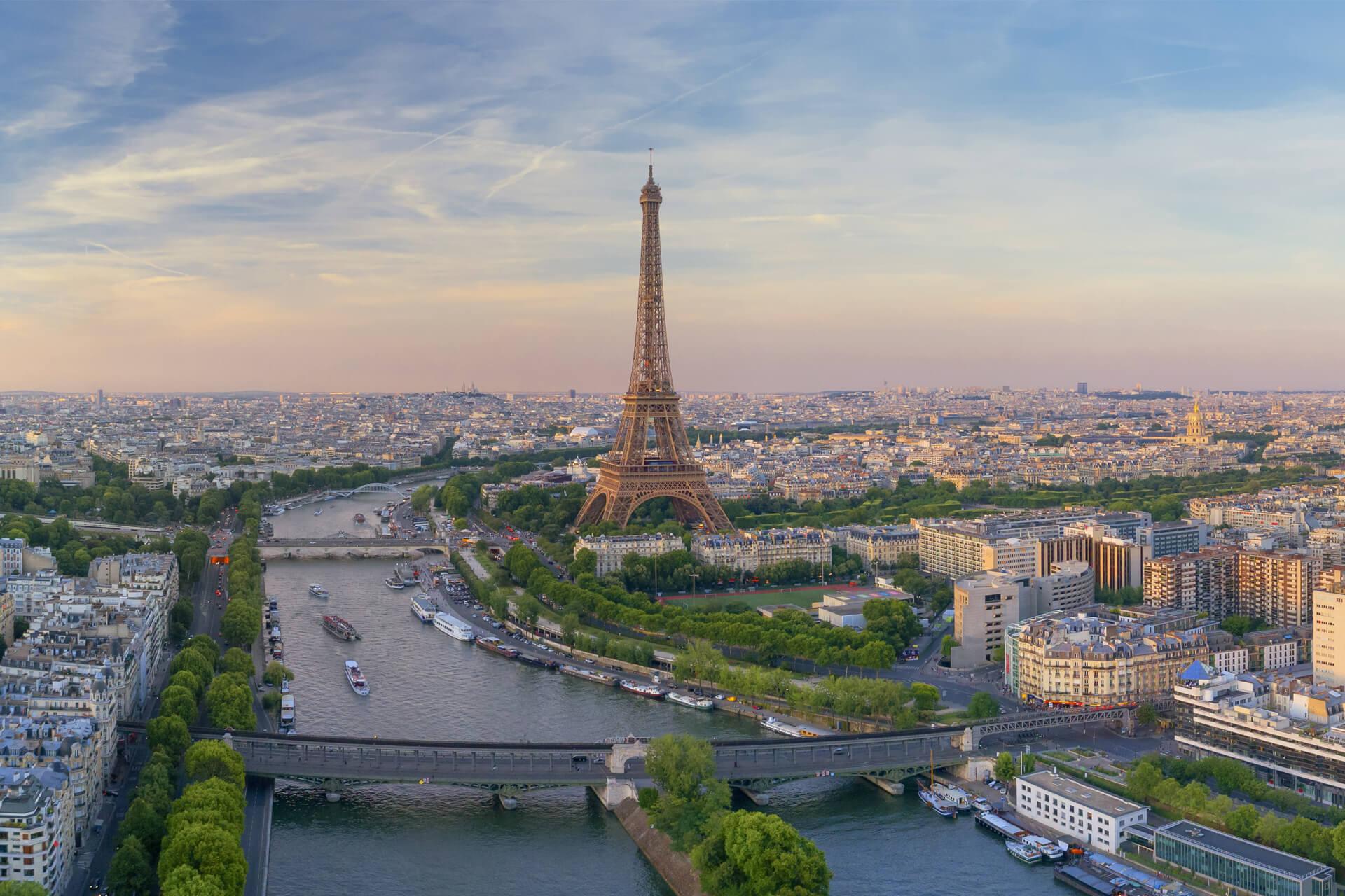 Vue aérienne panoramique de Paris au coucher du soleil avec la tour Eiffel au centre et des bateaux-mouches sur la Seine.