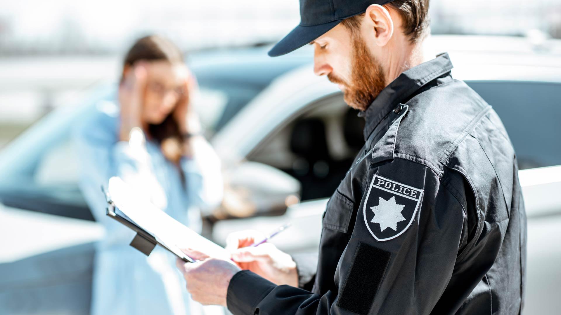 Close-up of a police officer filling out a report. Behind him, blurred, a woman leaning on her vehicle holds her head in her hands.