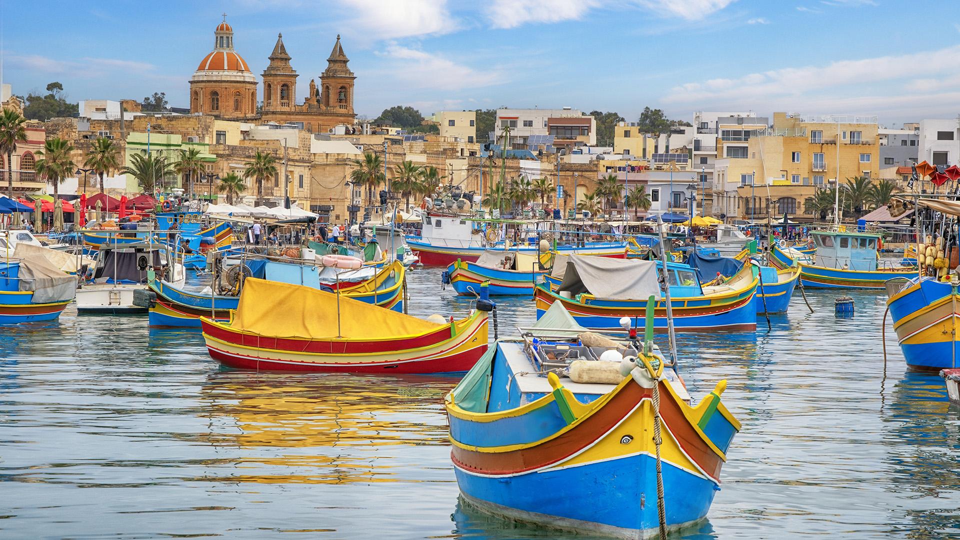 Port de Marsaxlokk à Malte, avec barques traditionnelles colorées, appelées luzzu, sur une eau calme et village en arrière-plan.
