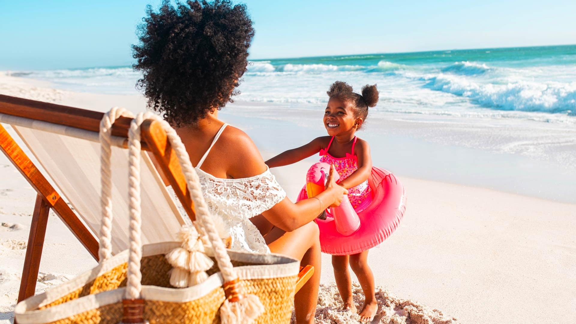 Sur une plage, une fillette portant maillot et flotteur de pélican rose tient les mains de sa mère assise sur une chaise.