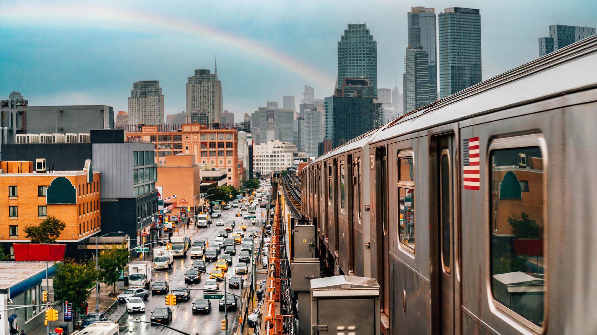 Métro aérien du Queens, à New York, surplombant une avenue avec voitures et taxis jaunes, sous un ciel nuageux et un arc-en-ciel.