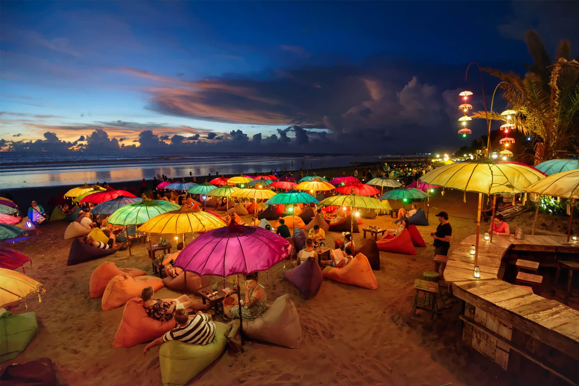 Parasols colorés et illuminés sur une plage animée de Seminyak en soirée avec des gens qui se détendent sur des poufs.