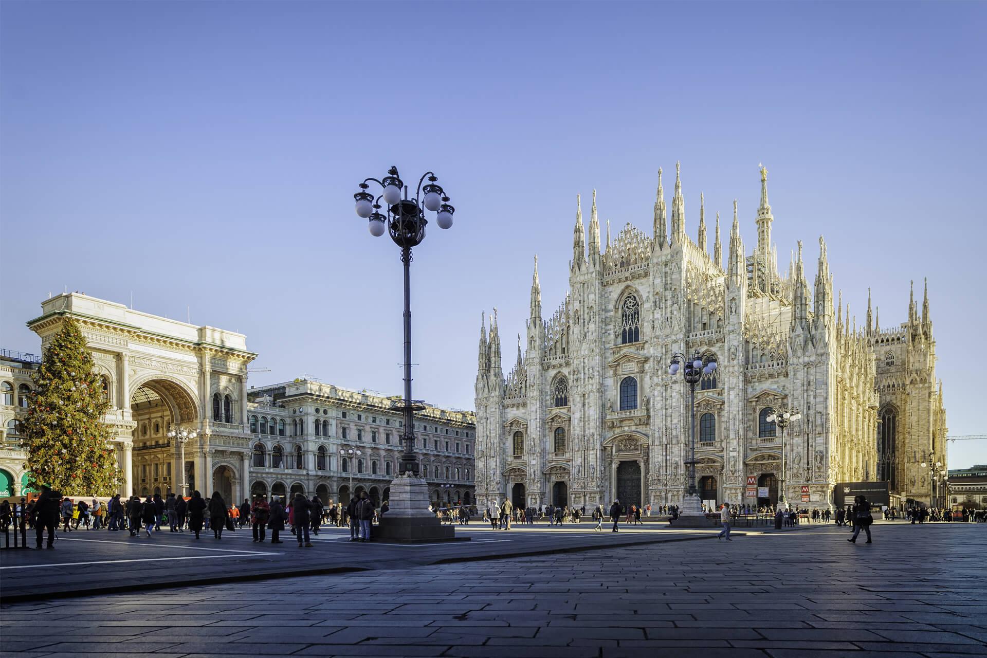 Piazza del Duomo in Milan, Italy, with the Duomo di Milano Cathedral and the Galleria Vittorio Emanuele II illuminated under a blue sky.