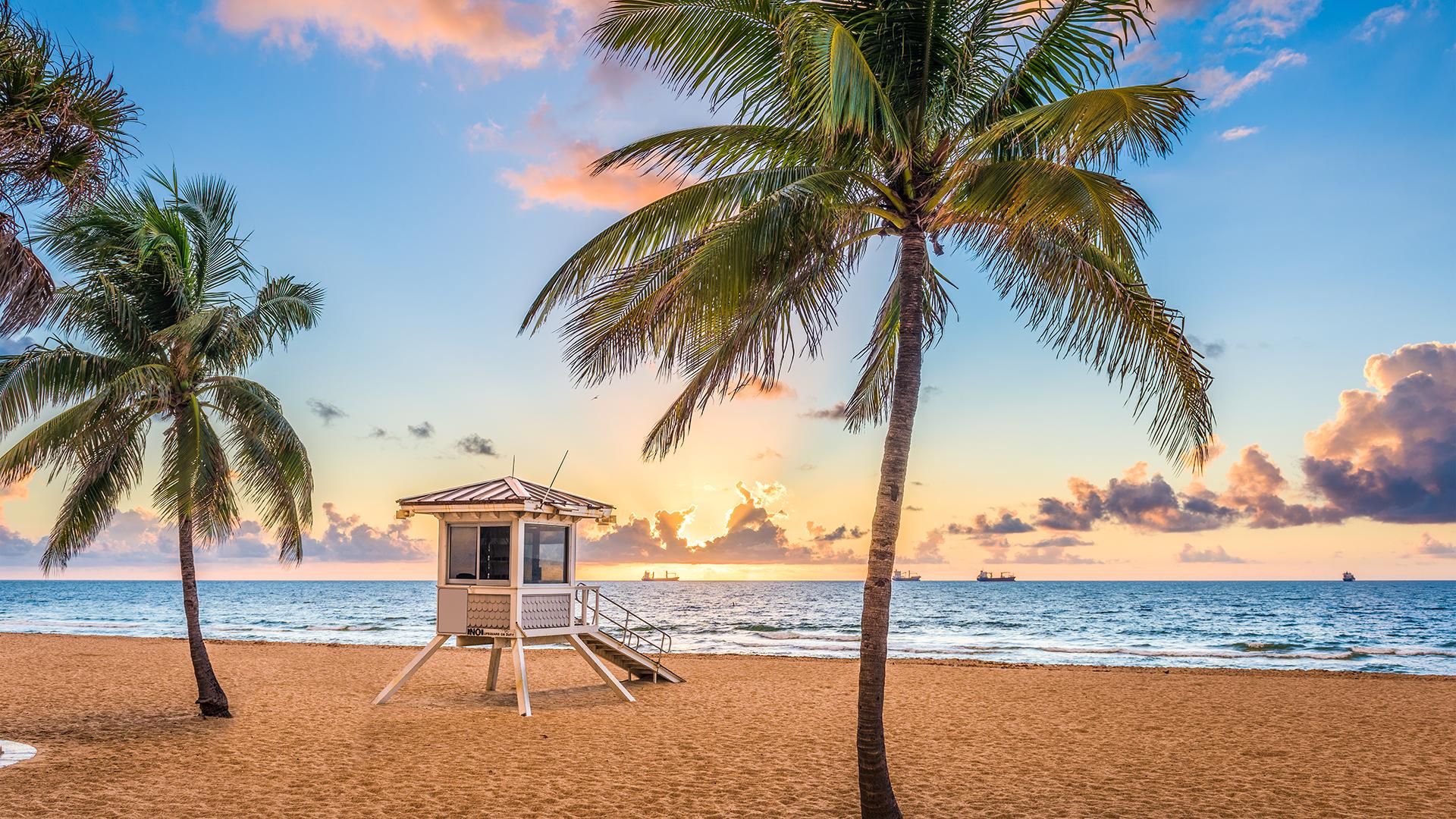 Lever du soleil sur une plage de Fort Lauderdale en Floride avec palmiers, cabane de sauveteur blanche et bateaux au loin.