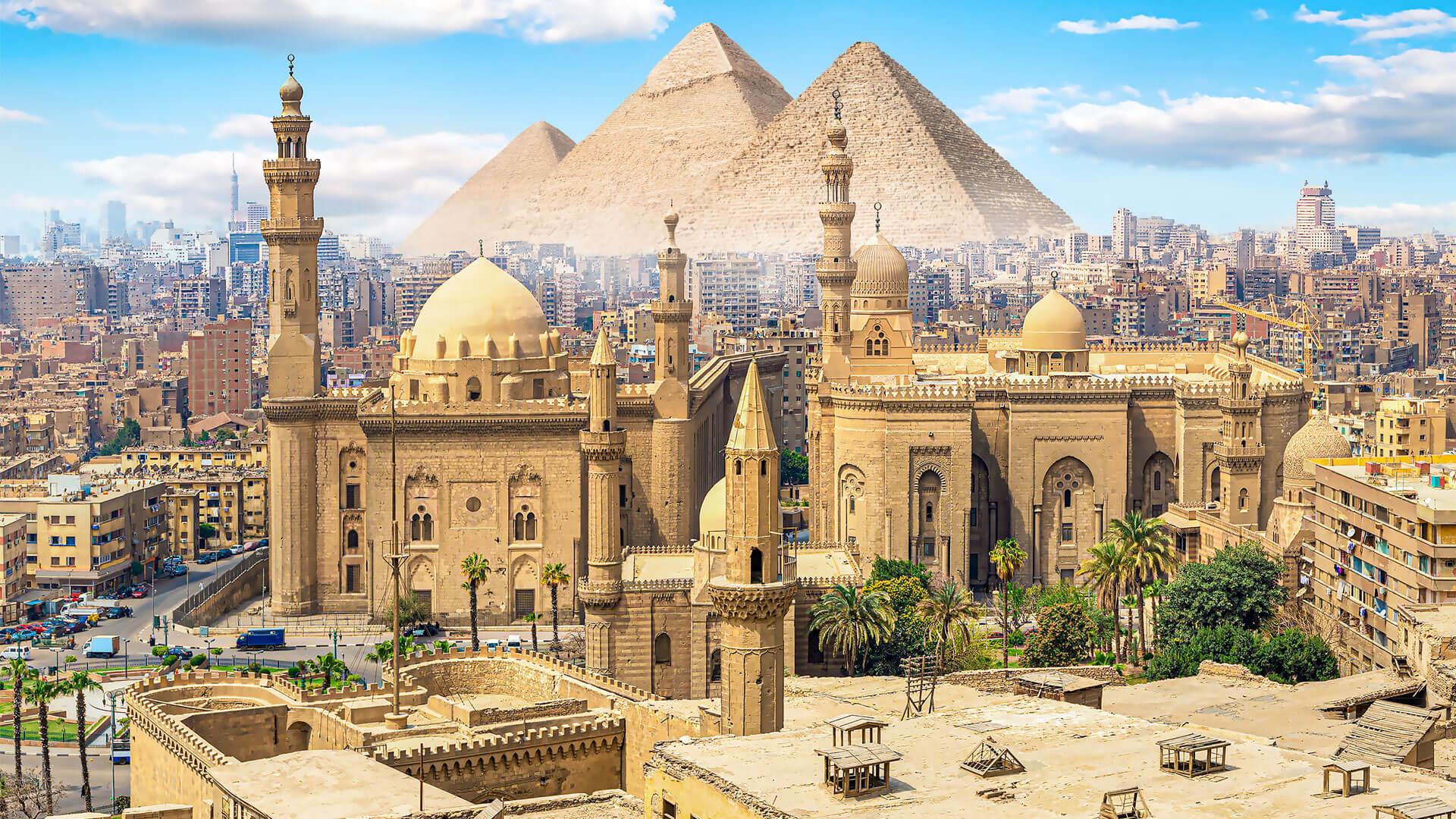 Vue du Caire avec mosquées historiques au premier plan et pyramides de Gizeh dominant la ville à l’horizon sous un ciel bleu.