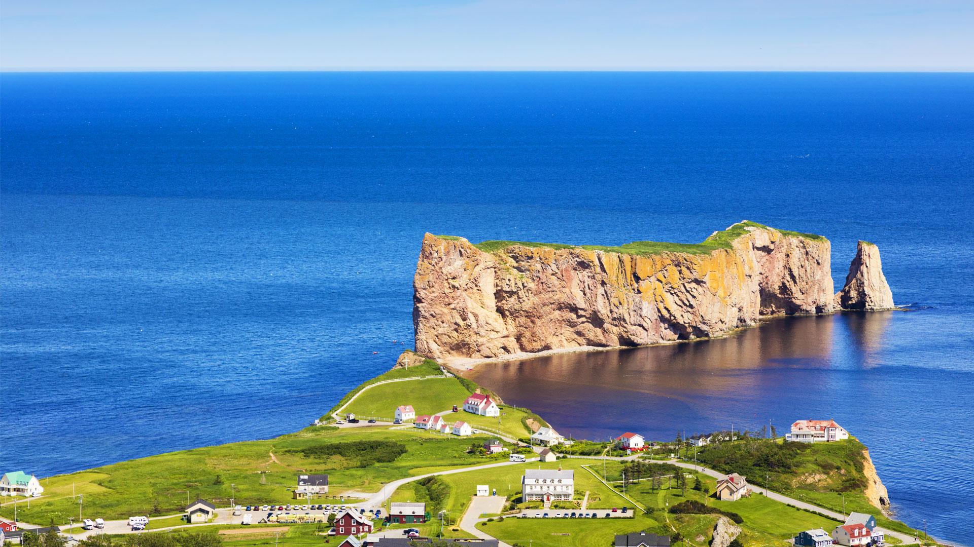 Aerial view of Percé Rock in the St. Lawrence Gulf, with the village of Percé in Gaspesie under a clear blue sky.