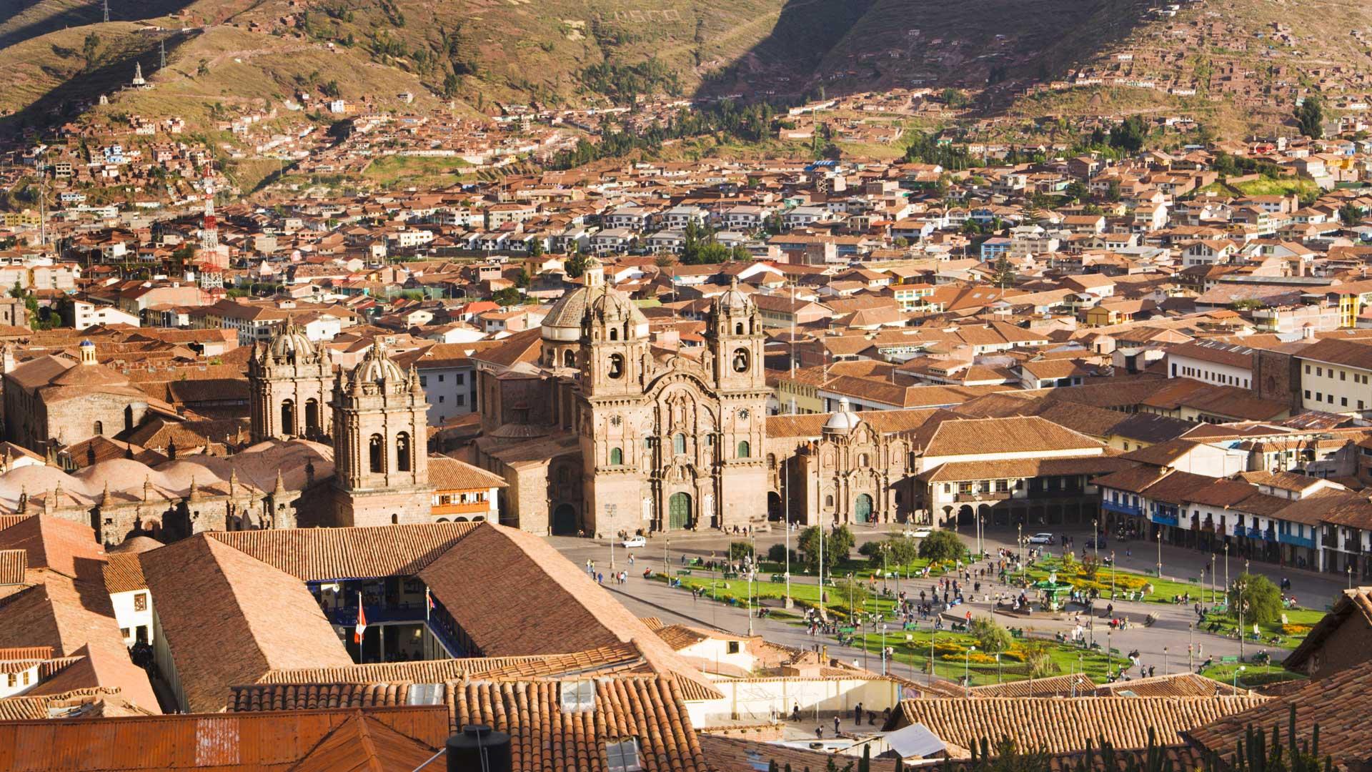 Panoramic view of Cusco, Peru, featuring the Plaza de Armas, colonial buildings with red-tiled roofs, and the Andes Mountains in the background.