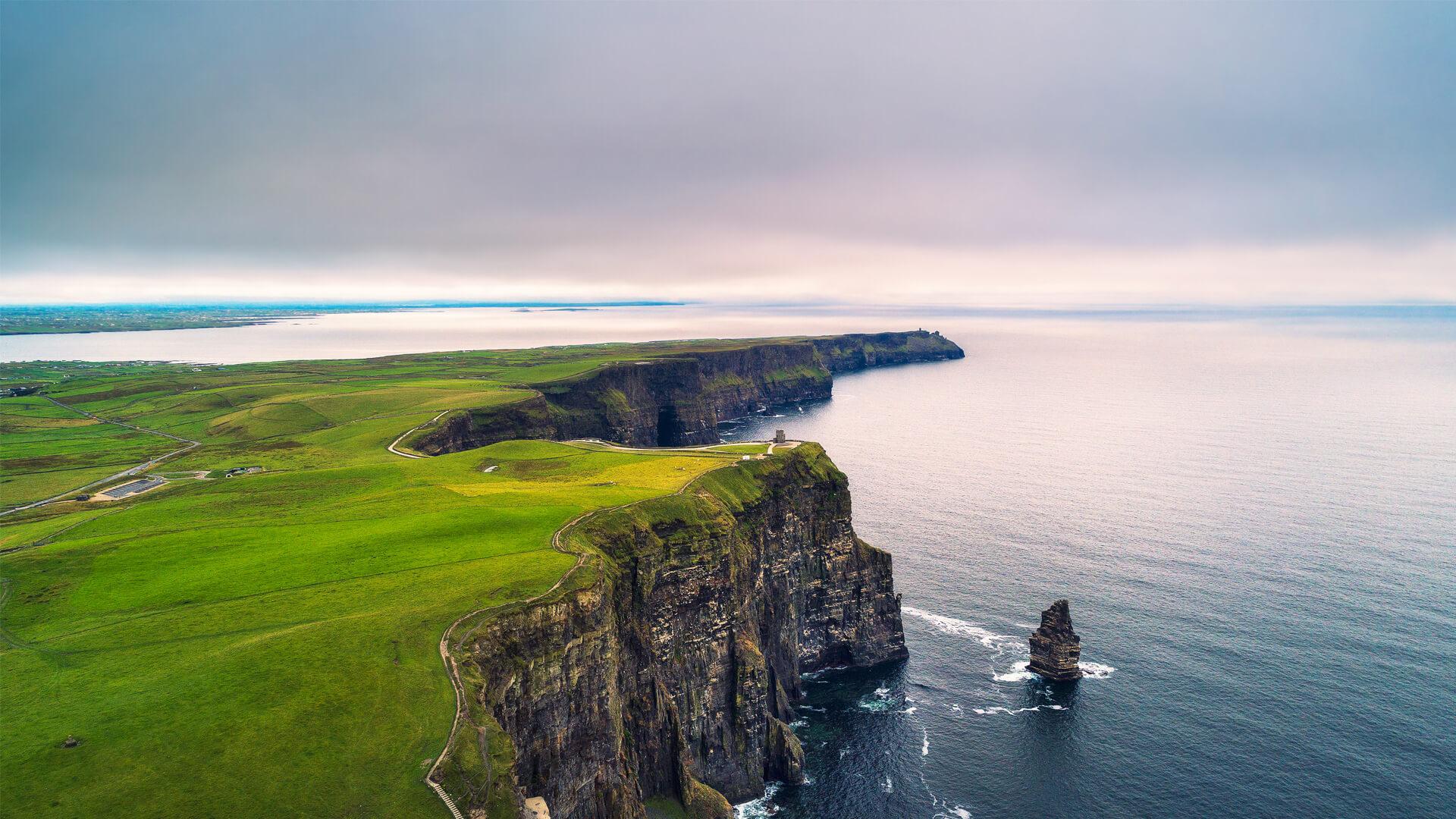 Vue aérienne des falaises de Moher en Irlande surplombant l’océan Atlantique, avec collines verdoyantes et ciel nuageux.