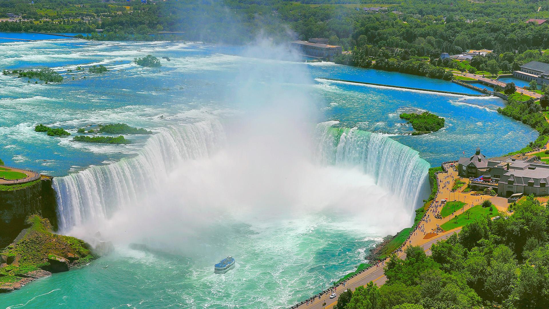 Vue aérienne des chutes du Niagara en Ontario avec un bateau d’excursion, de la brume et une végétation luxuriante sous un ciel bleu.